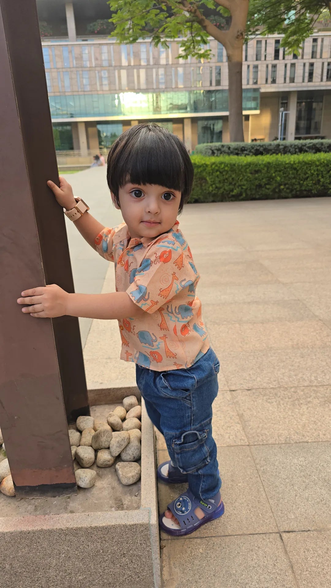 Child standing outdoors near a building with trees and pavement.