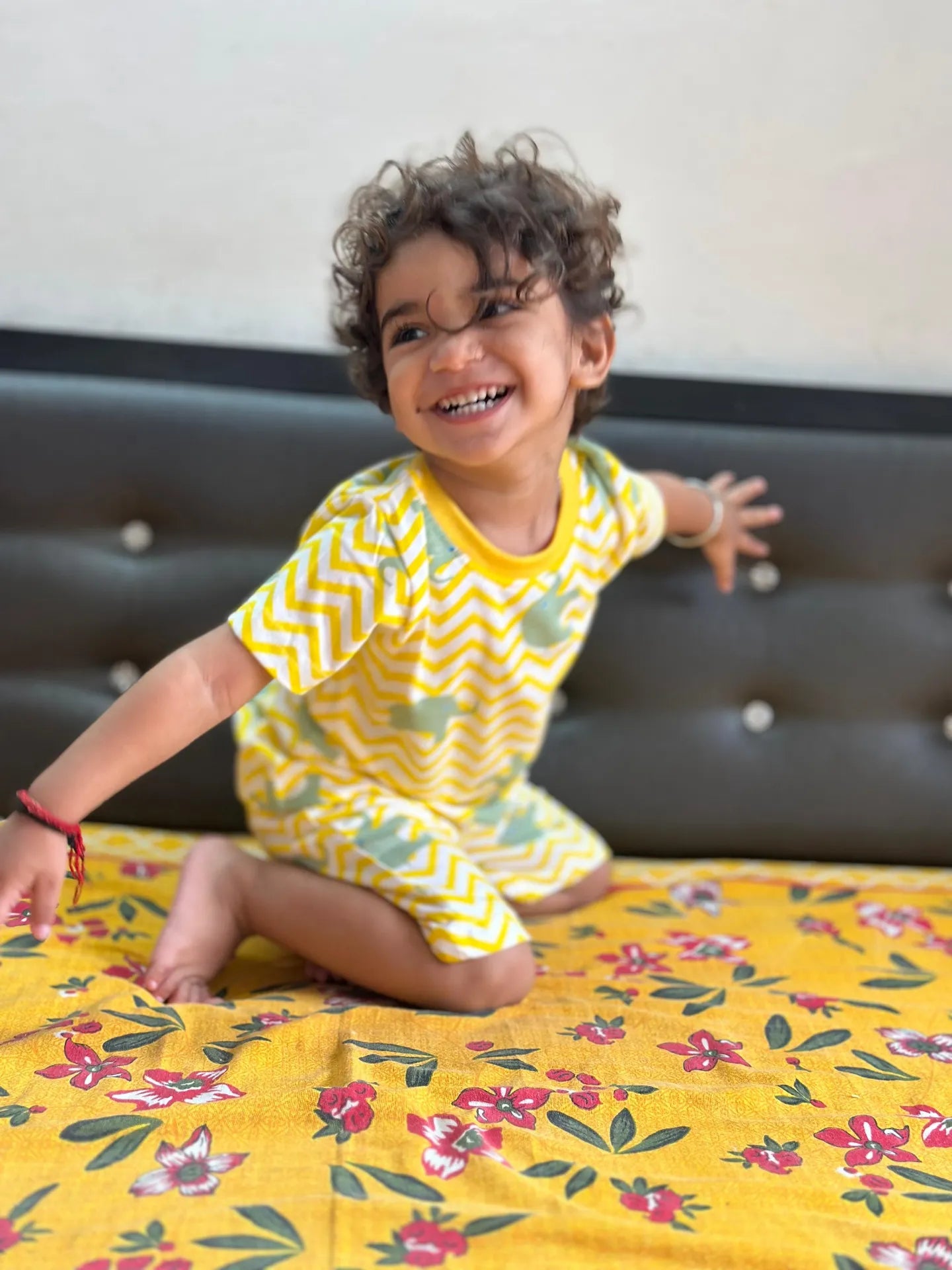 Child wearing a yellow and green outfit sitting on a floral bedspread.